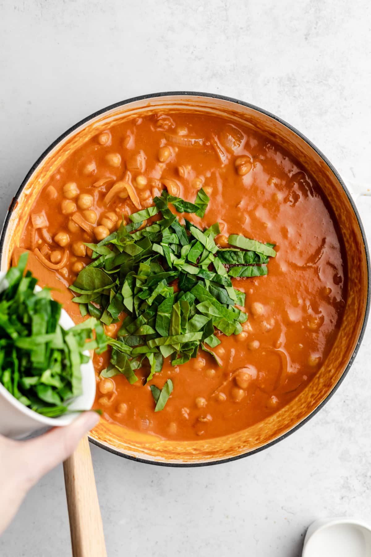 spinach being added to a pot of chickpeas and onions in a tomato sauce