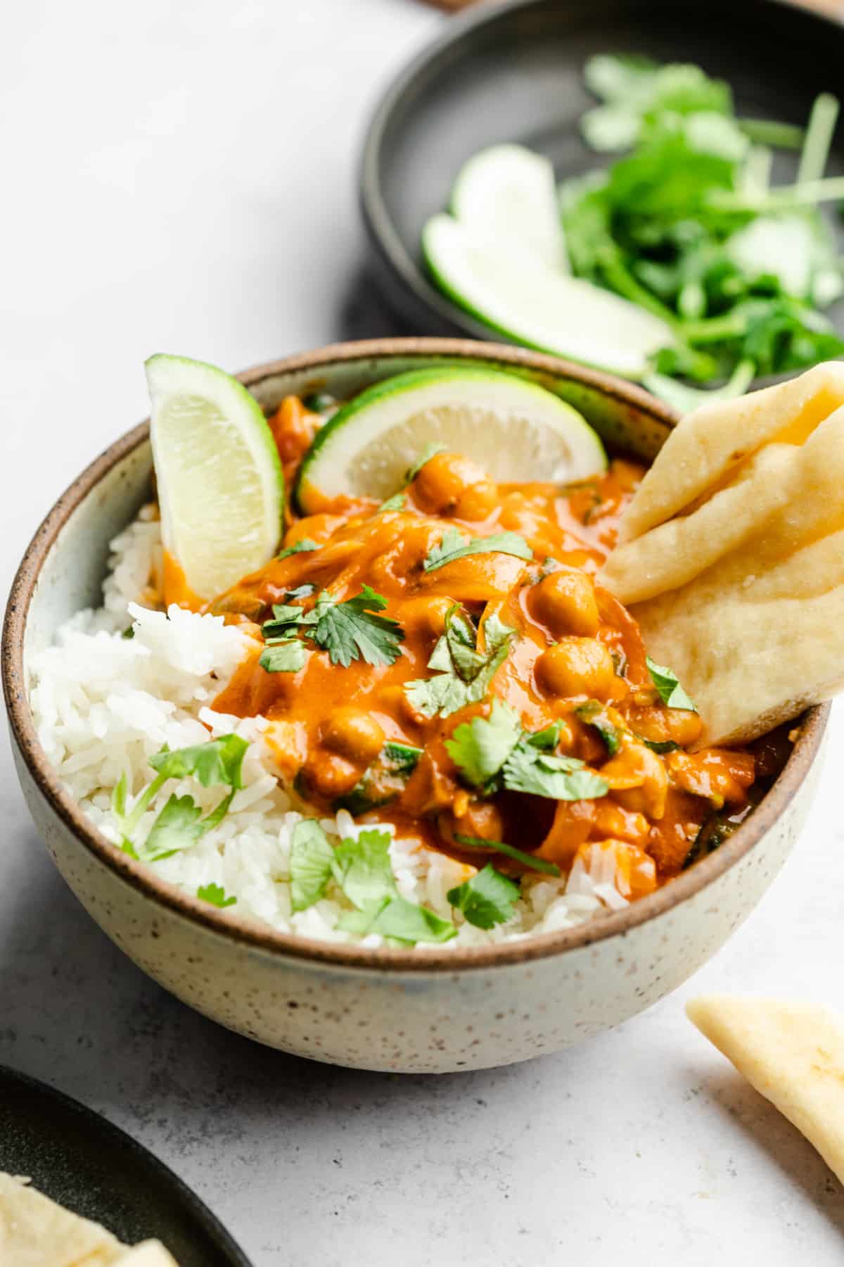 speckled bowl filled with rice, chickpea curry, cilantro, a lime and naan on greyish background