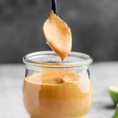 a spoon lifting up from a small glass jar of orange chipotle sauce.