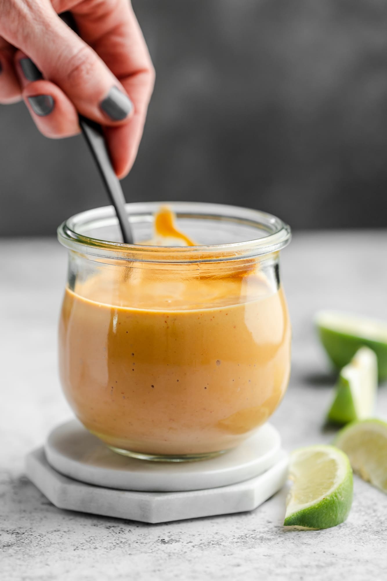 womans hand holding a spoon in a jar of orange chipotle sauce.