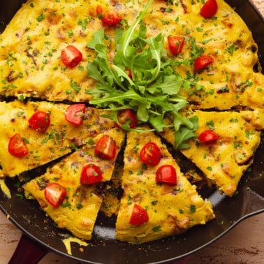 a cooked vegan frittata topped with sliced cherry tomatoes and arugula in a large black skillet.