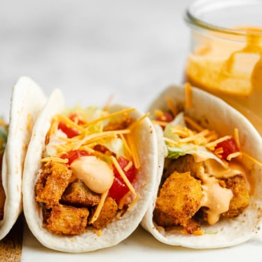 close up on 2 potato tacos next to each other topped with shredded cheddar cheese, chipotle sauce, lettuce, and tomatoes.