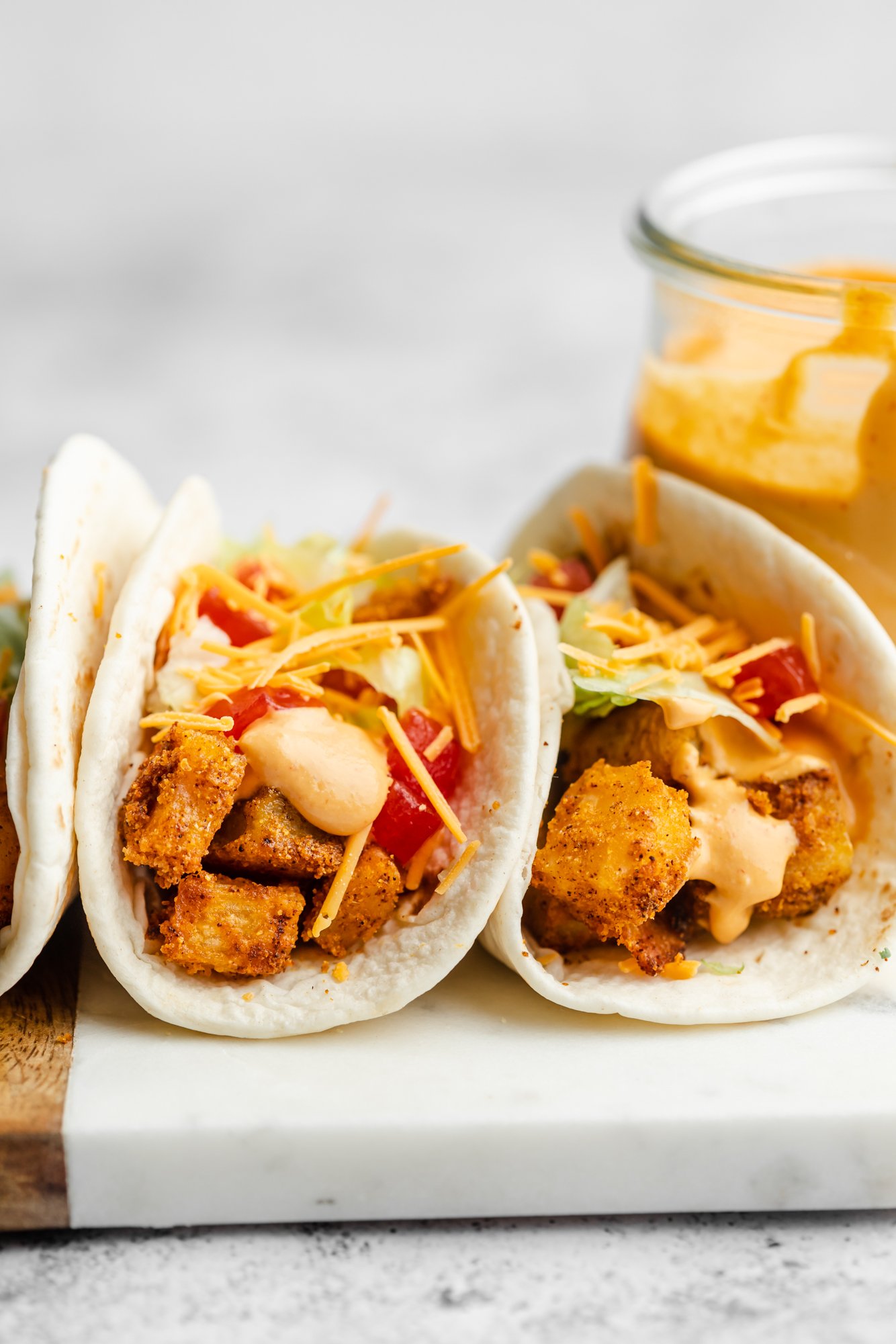 close up on 2 potato tacos next to each other topped with shredded cheddar cheese, chipotle sauce, lettuce, and tomatoes.