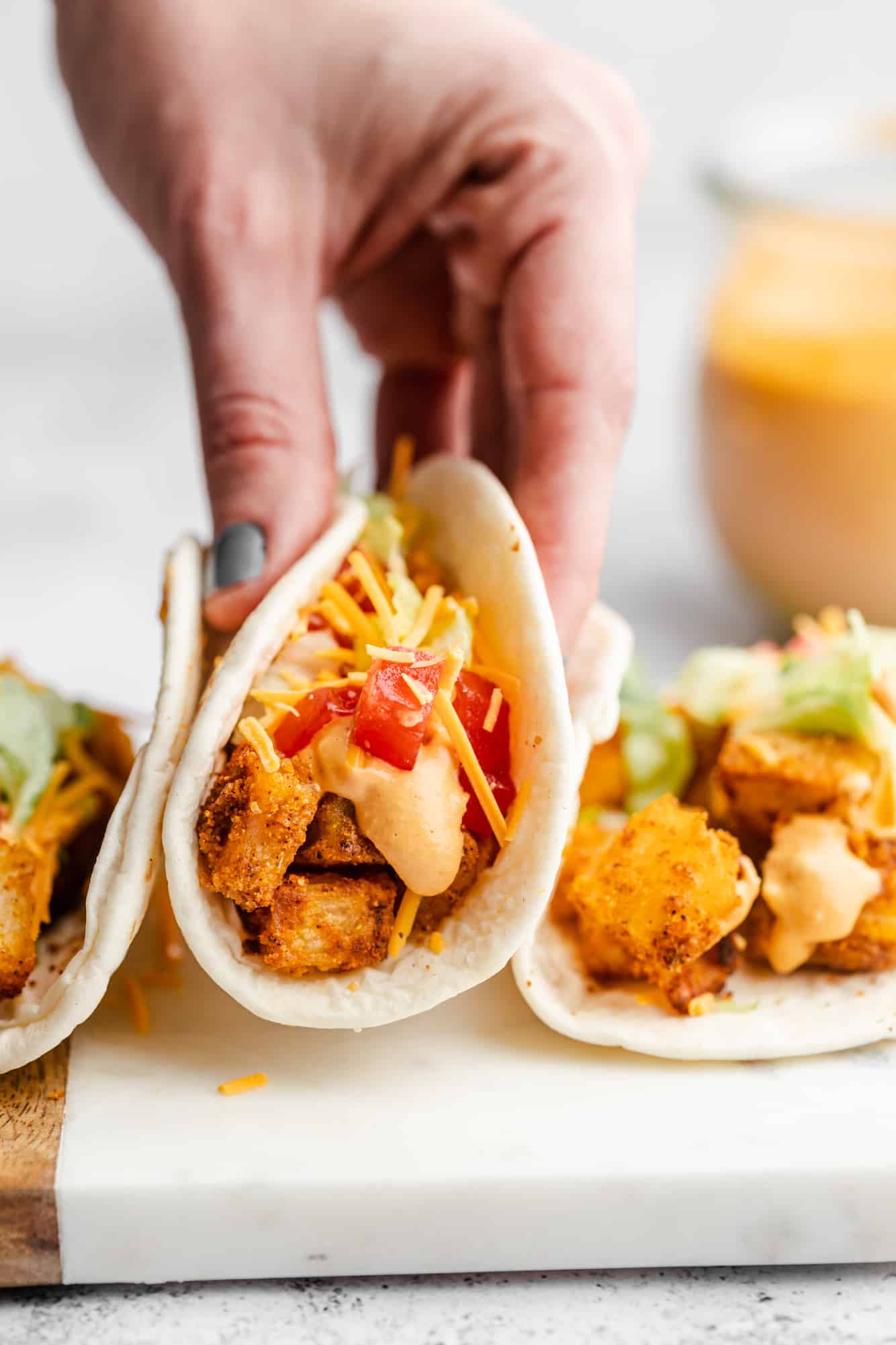 womans hand lifting a spicy potato taco from a row of other potato tacos.