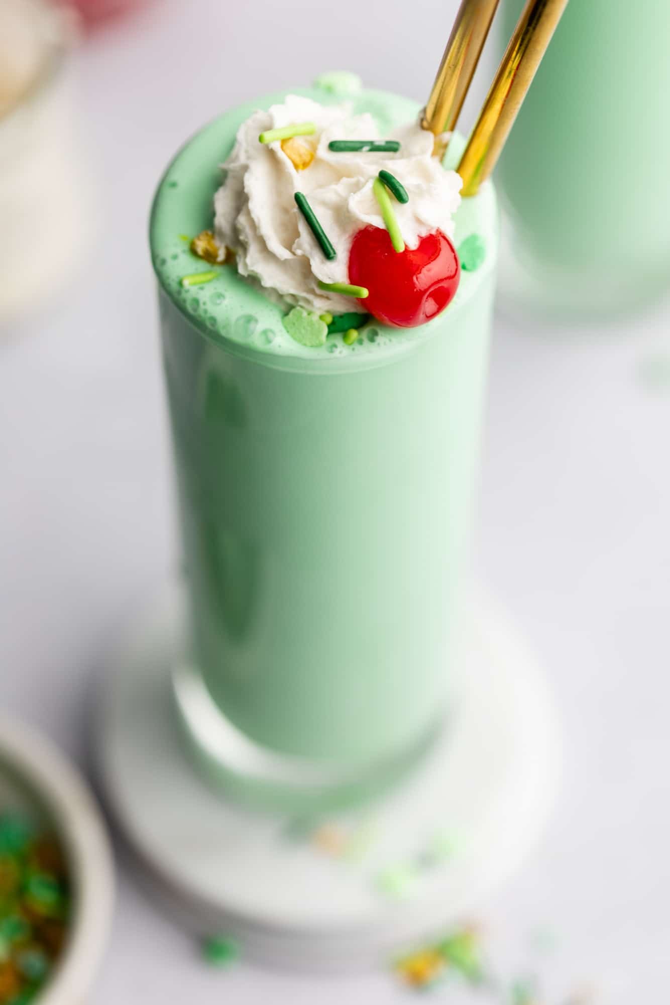 close up on a vegan shamrock shake in a tall glass topped with vegan whipped cream, green sprinkles, and a maraschino cherry.