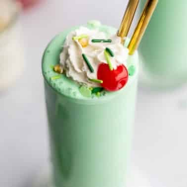 close up on a vegan shamrock shake in a tall glass topped with vegan whipped cream, green sprinkles, and a maraschino cherry.