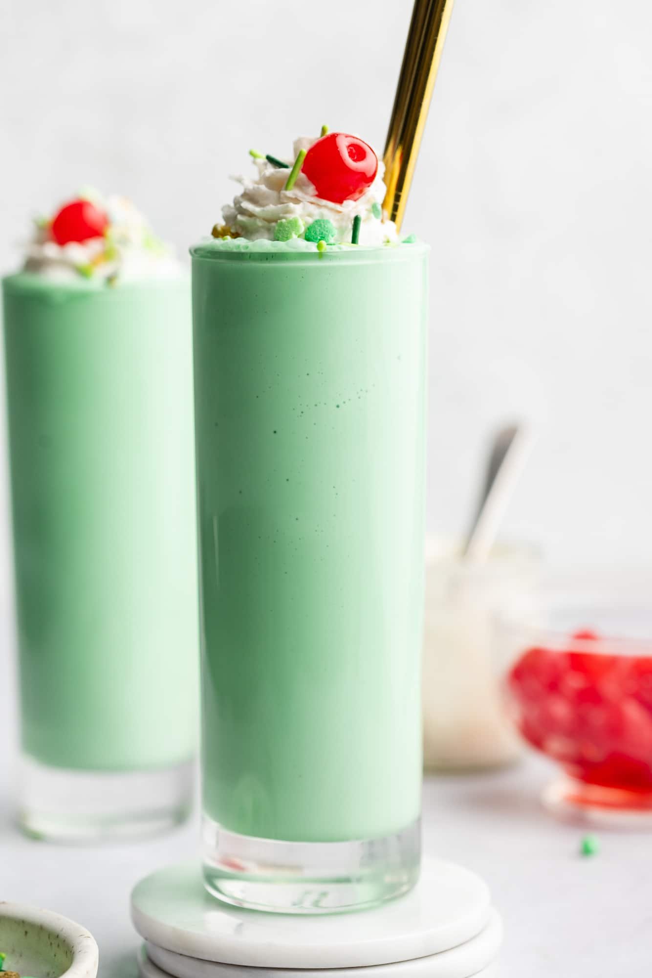2 mint green vegan shamrock milkshakes in tall glasses topped with whipped cream, green sprinkles, and a maraschino cherry.