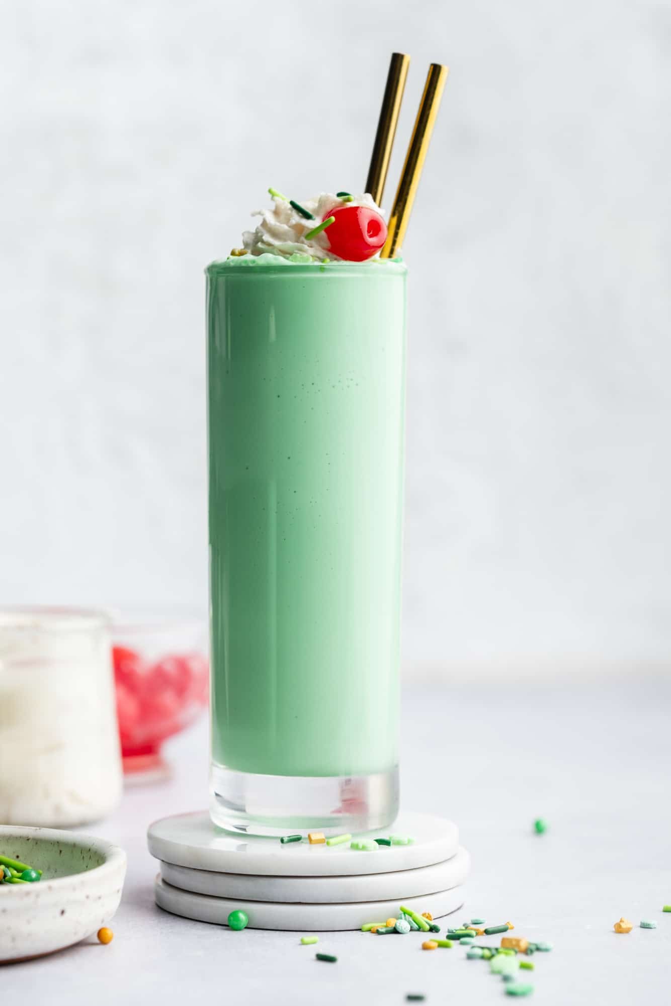 a vegan shamrock shake in a tall glass topped with vegan whipped cream, green sprinkles, and a maraschino cherry.