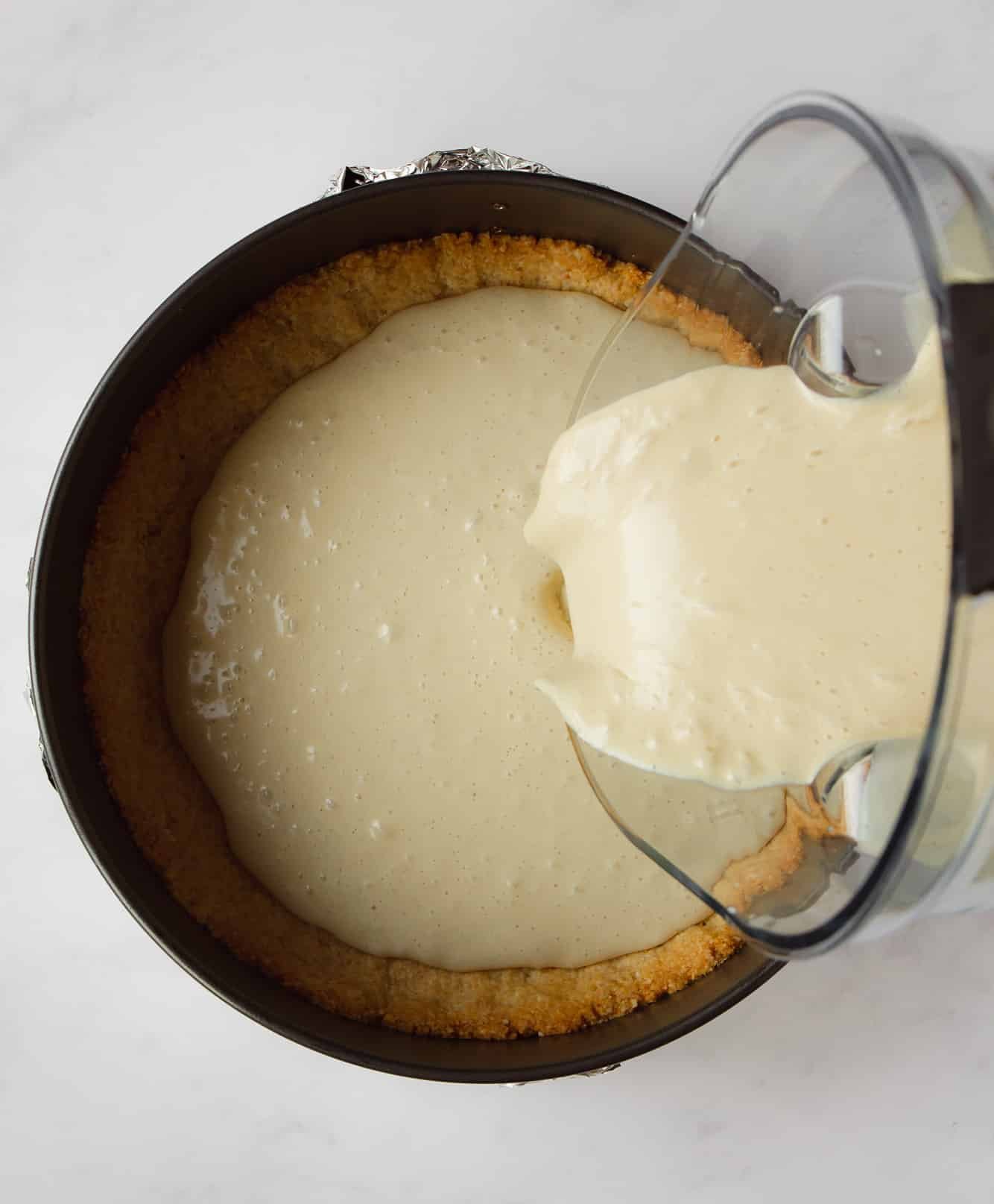 pouring a white sauce into a baked cheesecake crust.