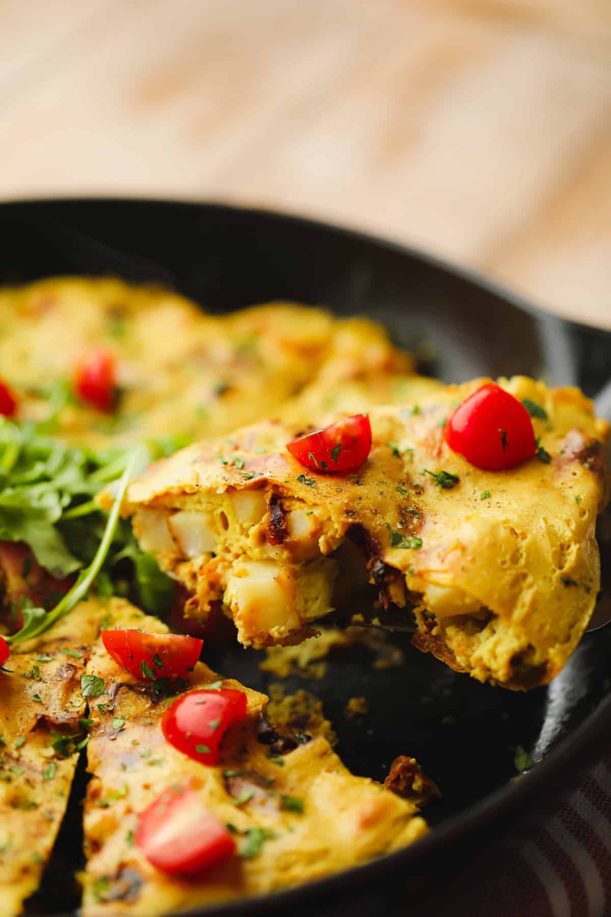 lifting a slice of vegan frittata topped with sliced cherry tomatoes from a black skillet.