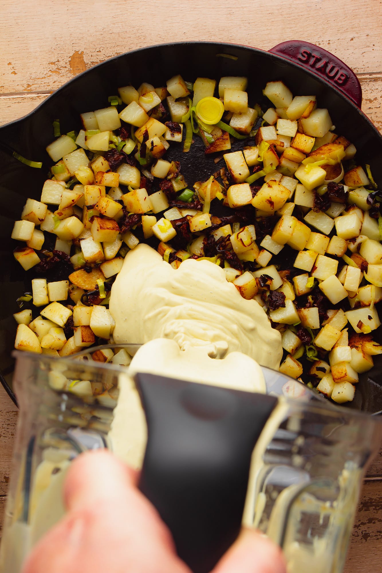 pouring a creamy yellow sauce over fried potatoes in a large black skillet.