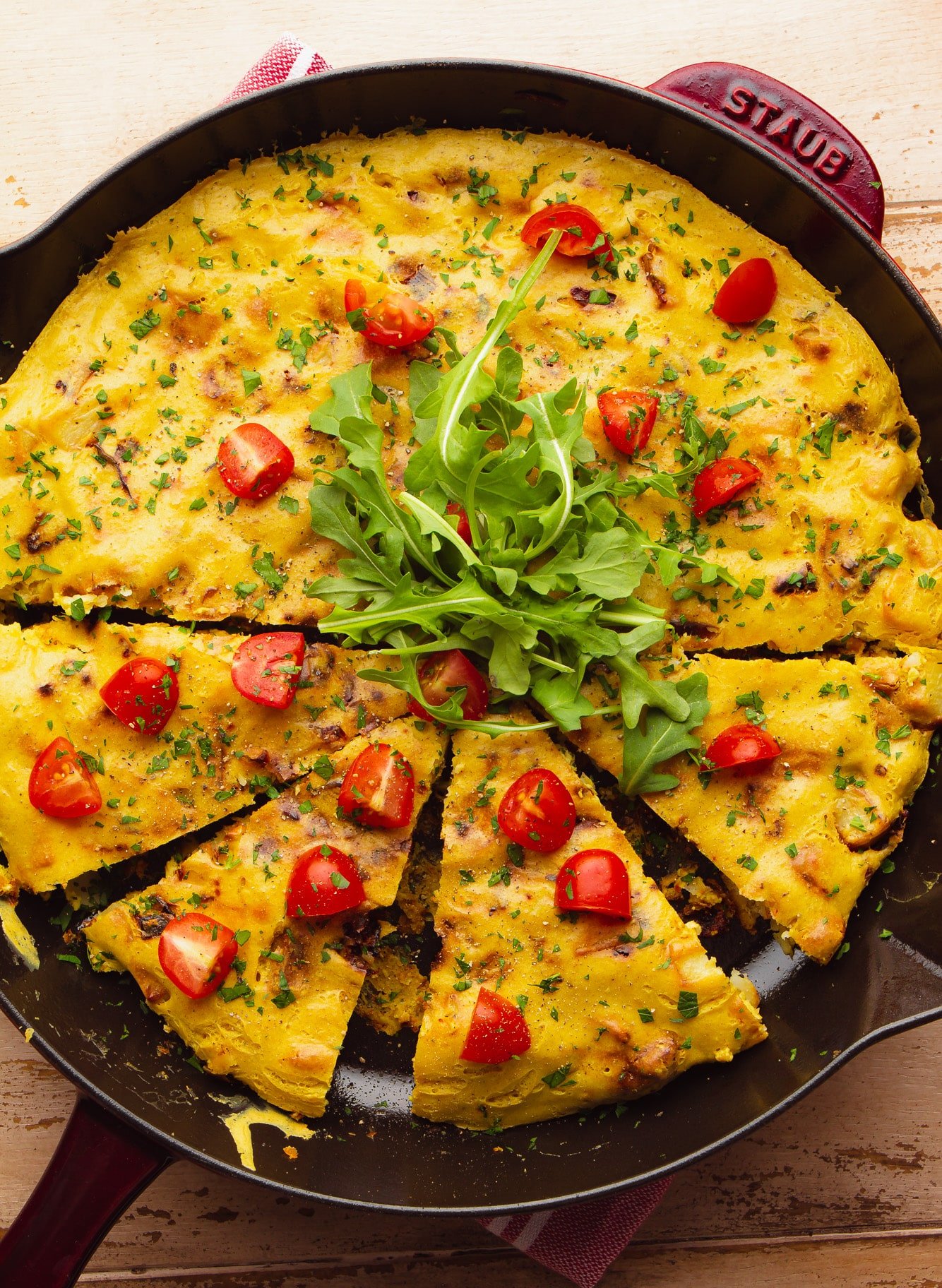 a cooked vegan frittata topped with sliced cherry tomatoes and arugula in a large black skillet.