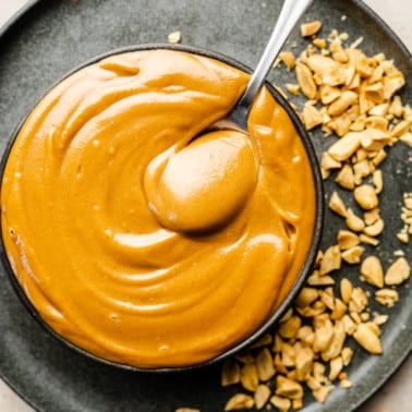 a spoon in a bowl of peanut sauce on top of a black plate with crushed peanuts.