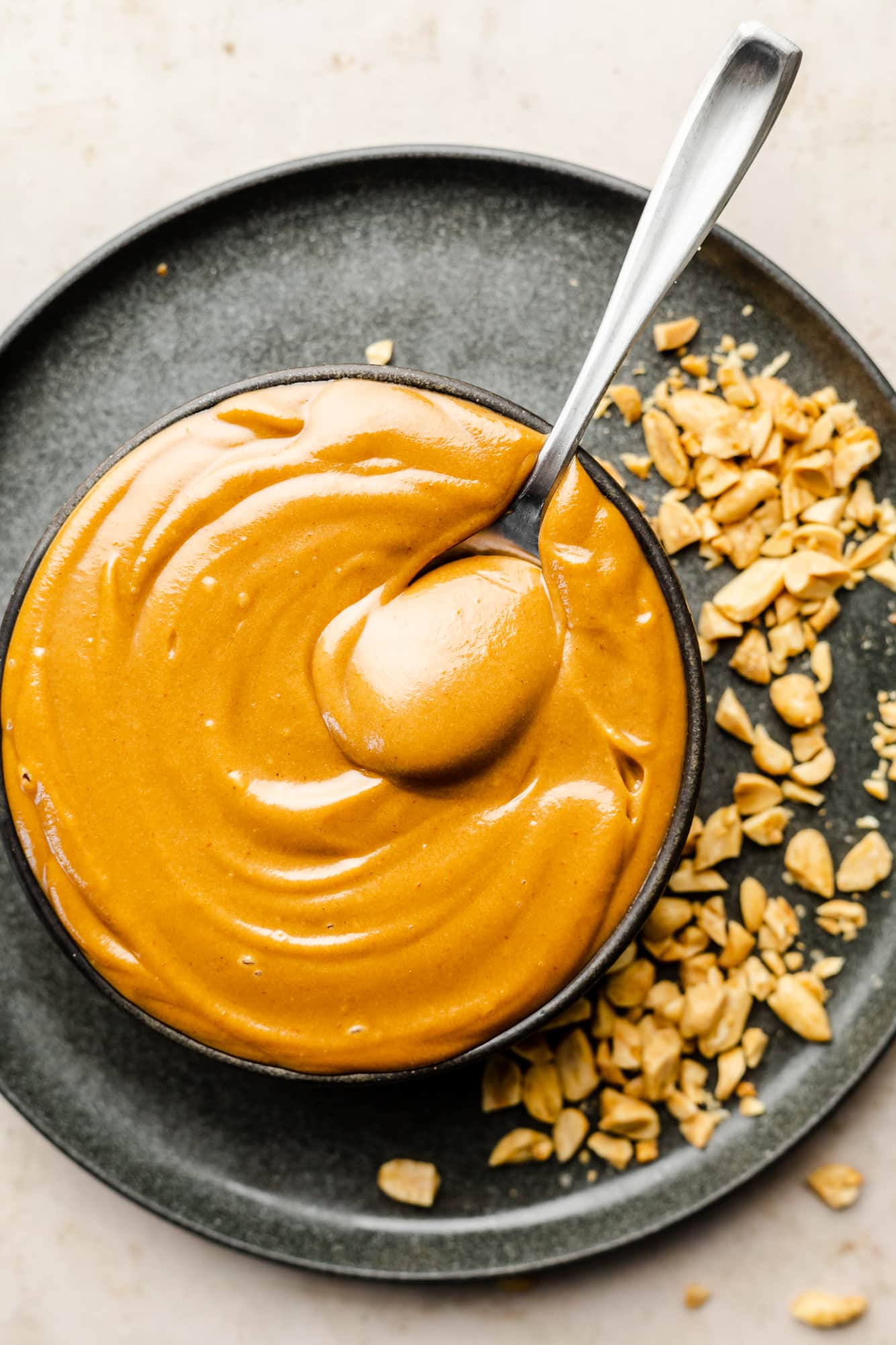 a spoon in a bowl of peanut sauce on top of a black plate with crushed peanuts.