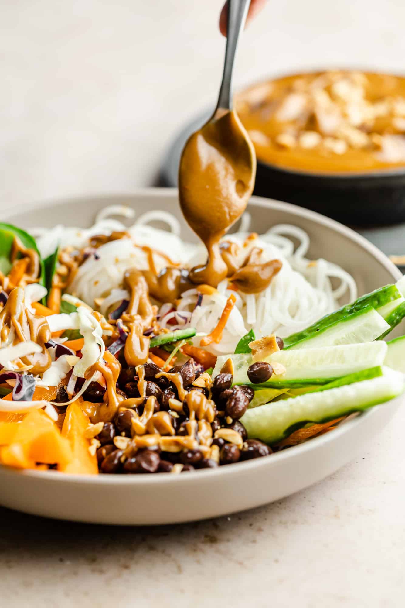 using a spoon to drizzle peanut sauce over a bowl of noodles and vegetables.