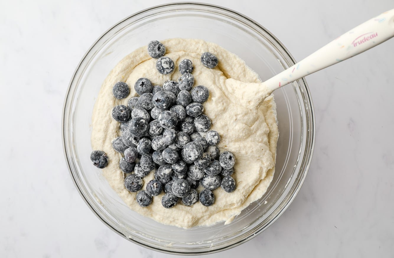 using a white spatula to fold blueberries into white cake batter.