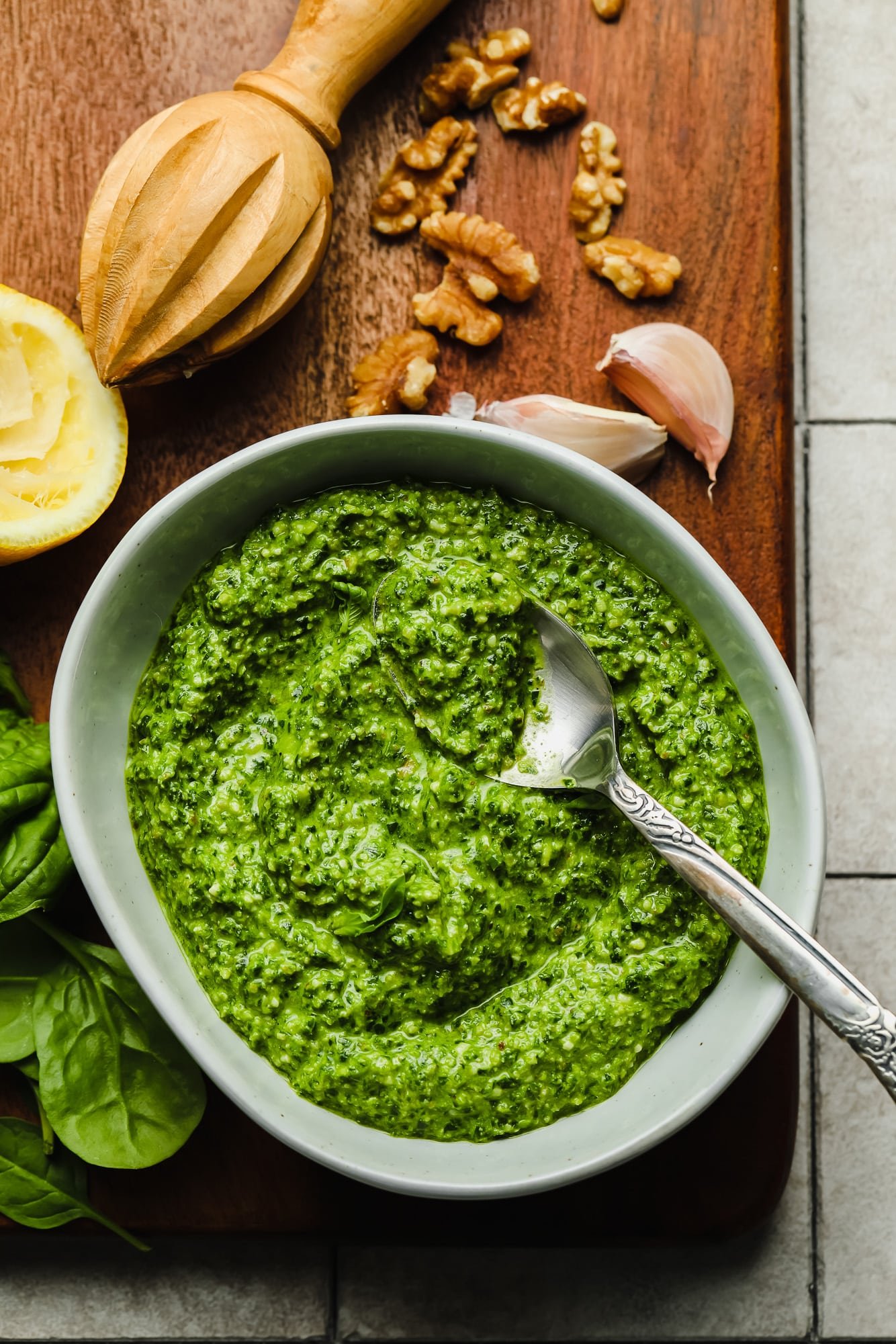 spinach pesto sauce in a grey bowl with a spoon on top, surrounded by nuts, garlic, spinach, and lemon.