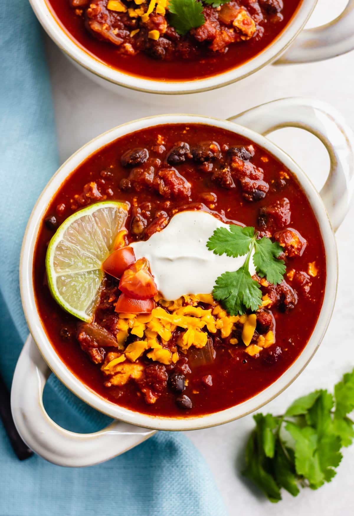 looking down on two bowls of chili with sour cream, cheese and a lime on white background with blue towel