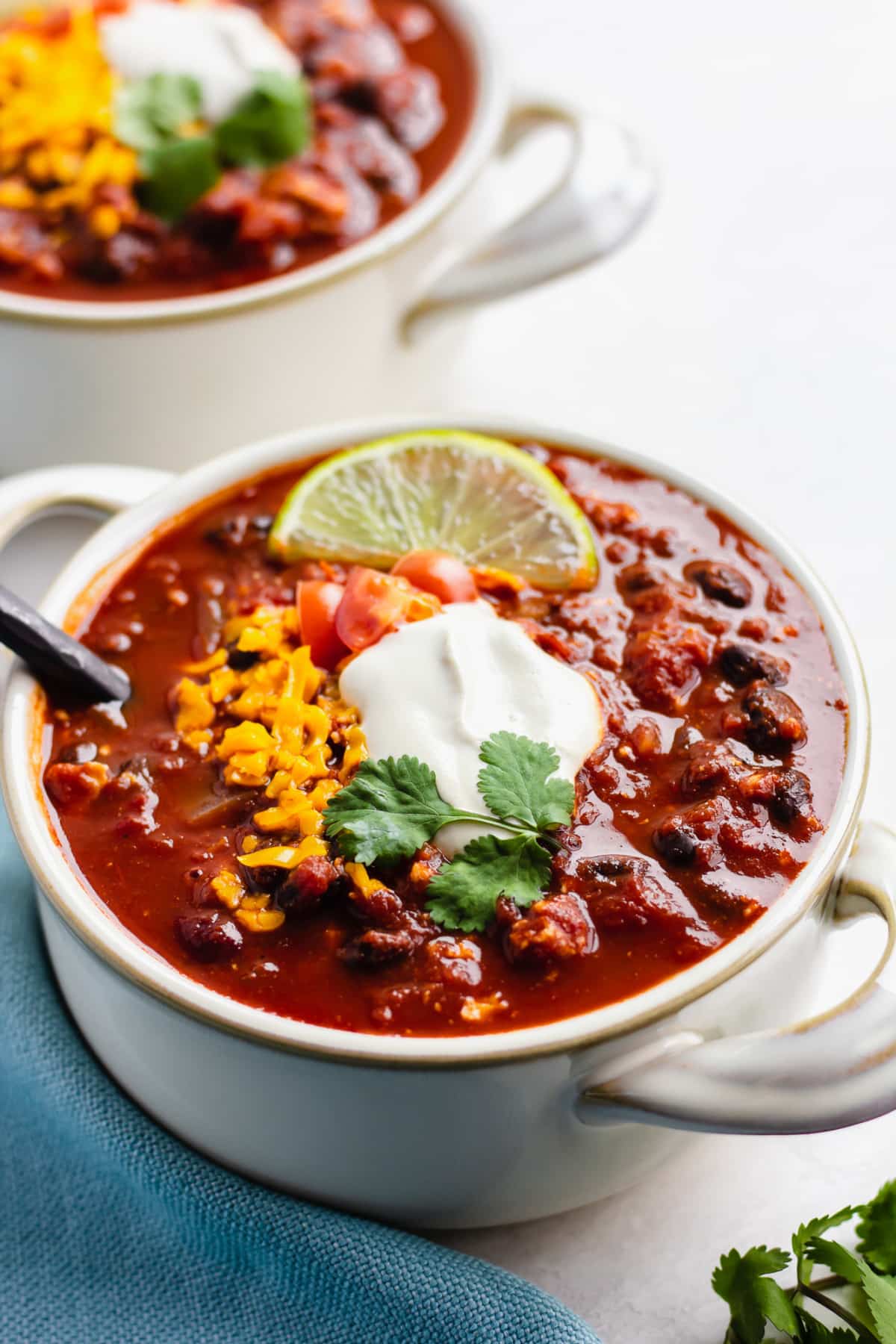 two bowls of vegan chili on white background with blue towel, sour cream and cheese on top.
