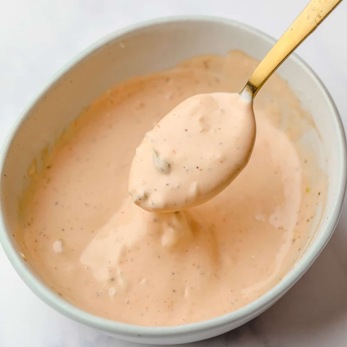 a gold spoon lifting up a scoop of vegan orange burger sauce from a white bowl.