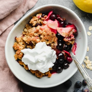 a serving of apple and blueberry crumble in a white bowl with vegan whipped cream on top.