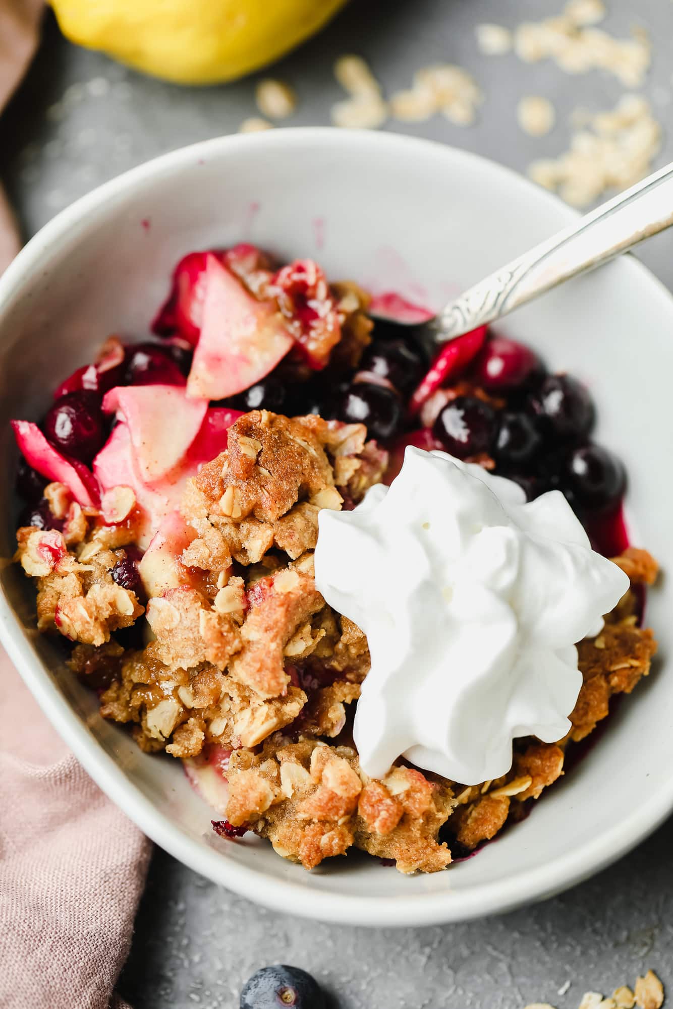close up on a serving of apple and blueberry crumble in a white bowl with vegan whipped cream on top.