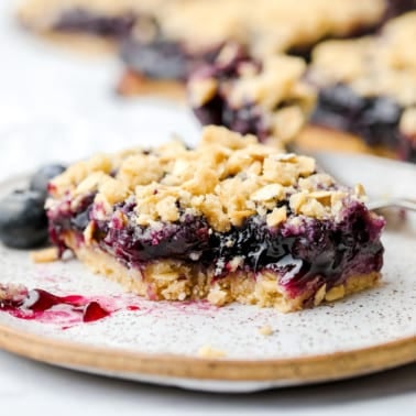 square image of a blueberry pie bar with oatmeal crumb topping, bite taken out of it on a speckled plate