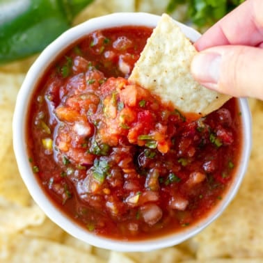 womans hand dipping a tortilla chip into a small bowl of tomato salsa.