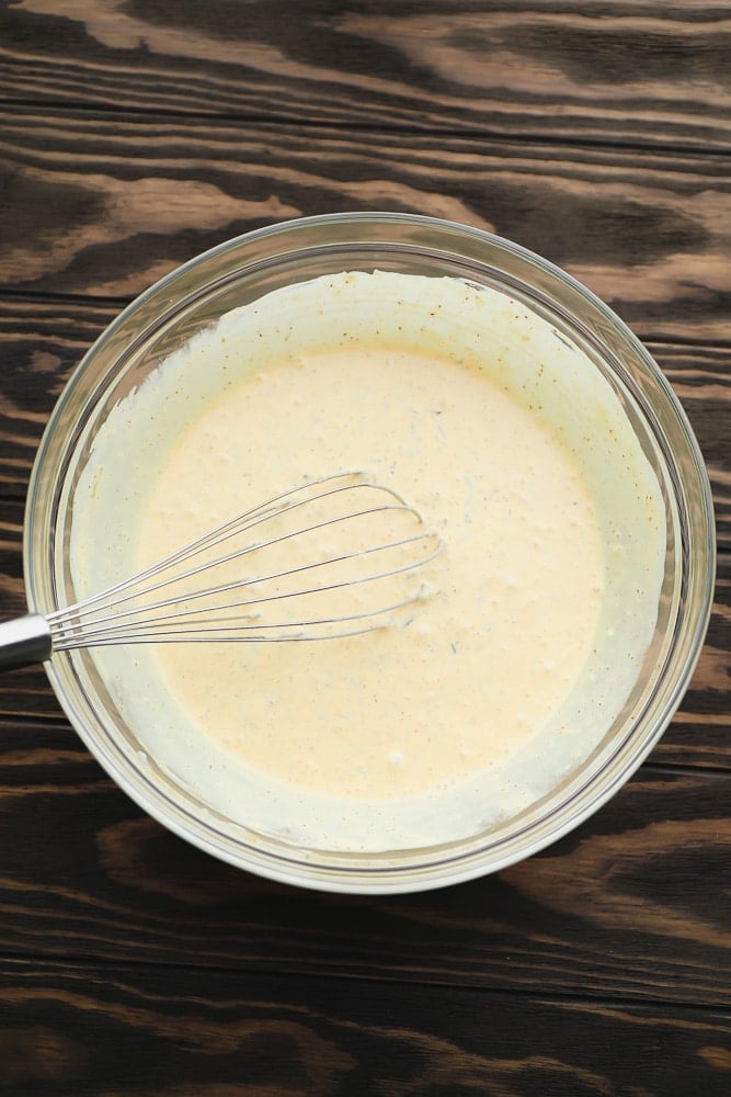 glass bowl with creamy yellow tinted dressing with a whisk in it on wood background