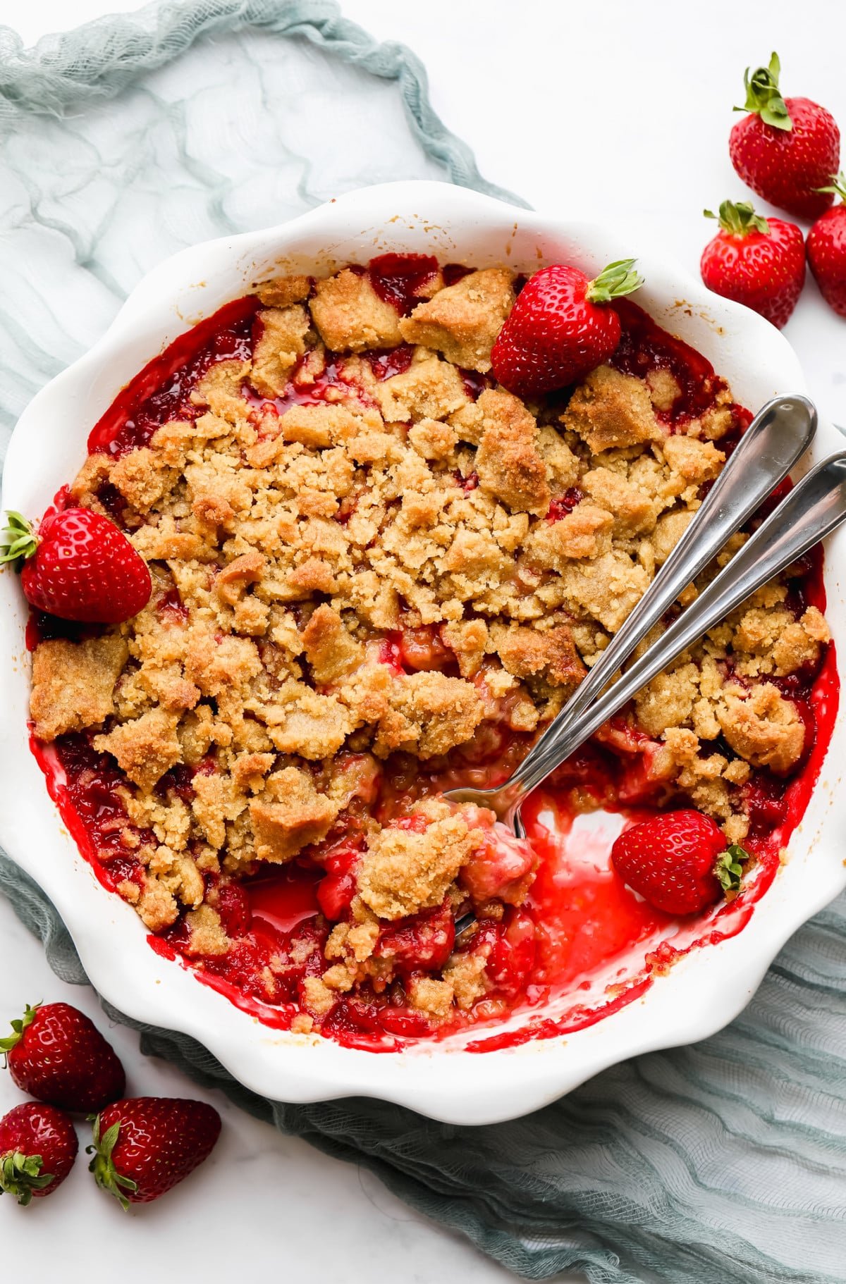 using metal spoons to scoop strawberry crumble from a white round dish.