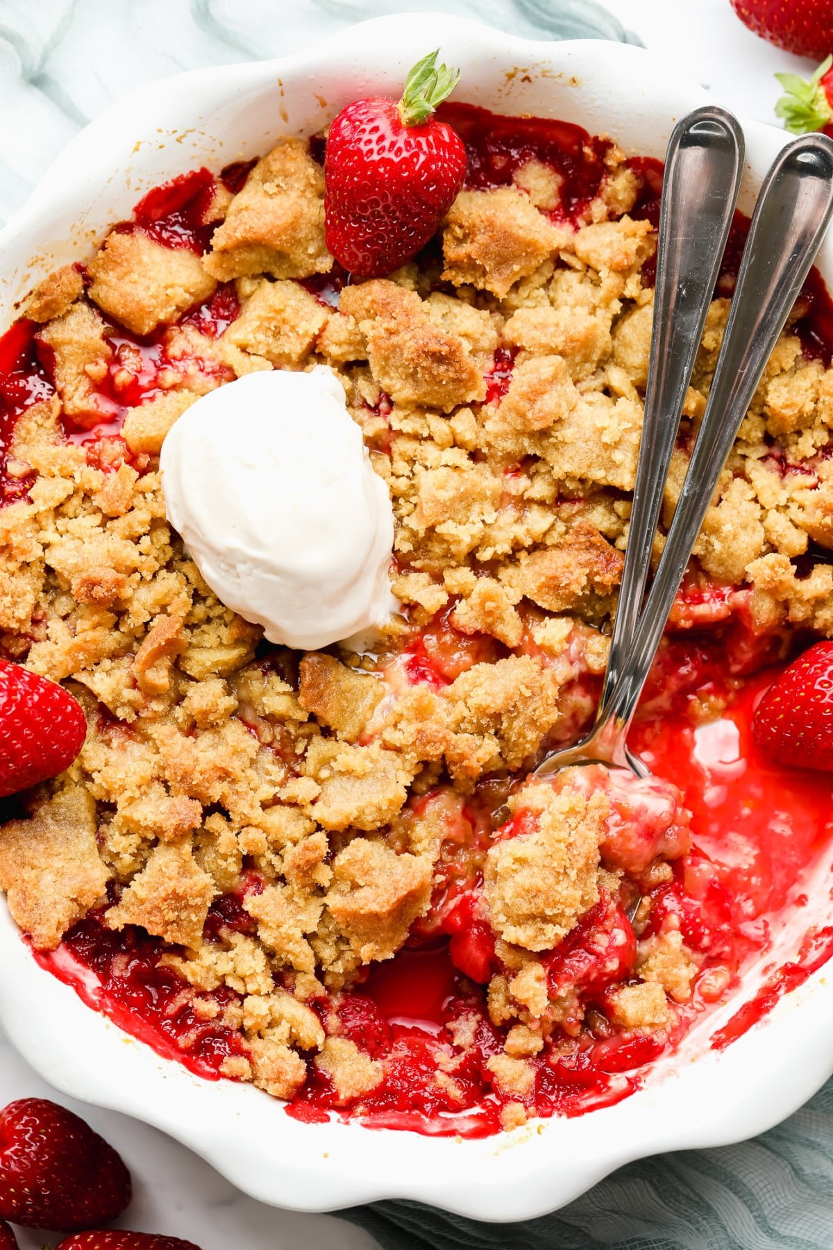 using metal spoons to scoop strawberry crumble from a white round dish.