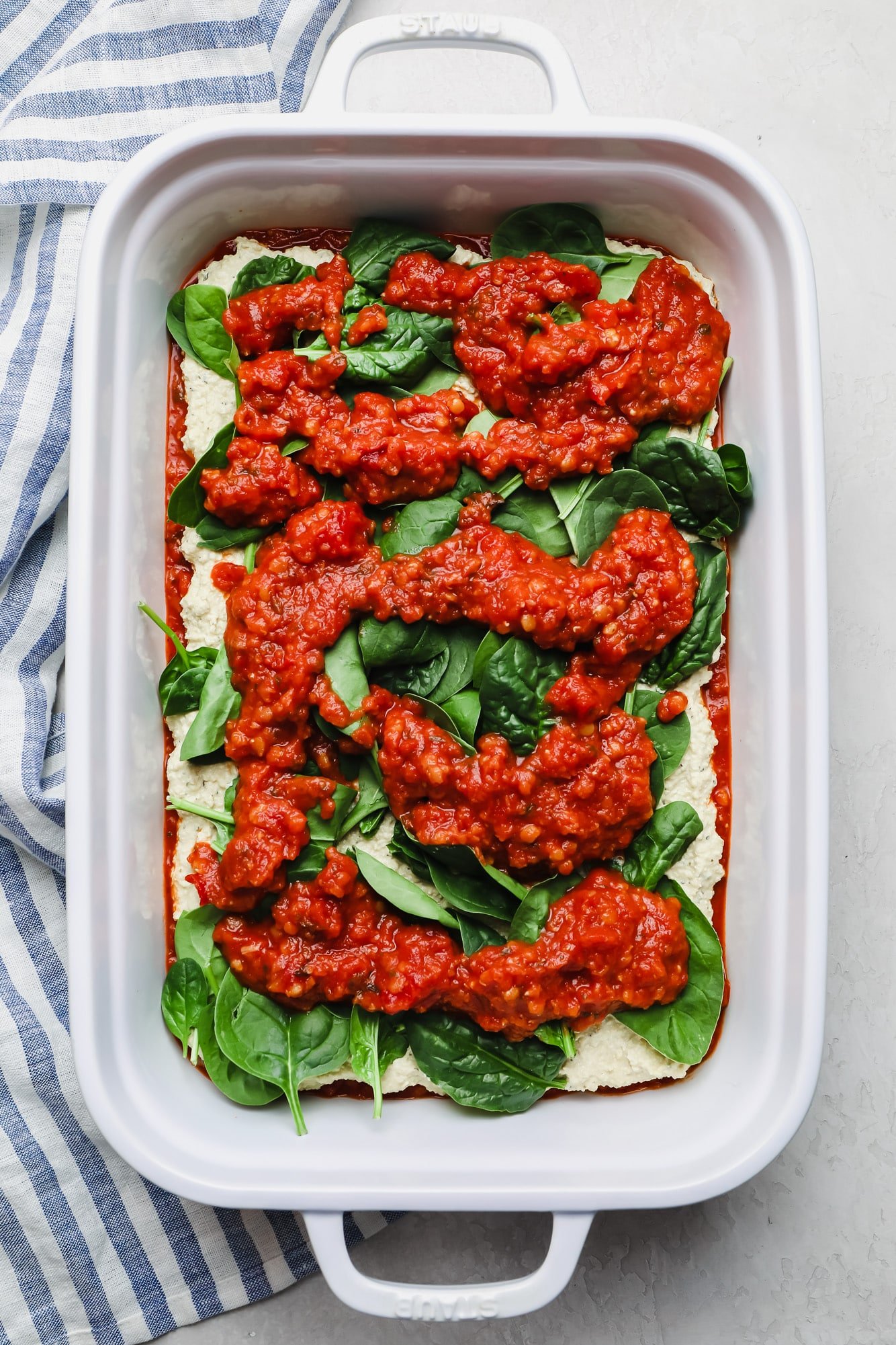 layering ingredients in a white casserole dish with red sauce and spinach.