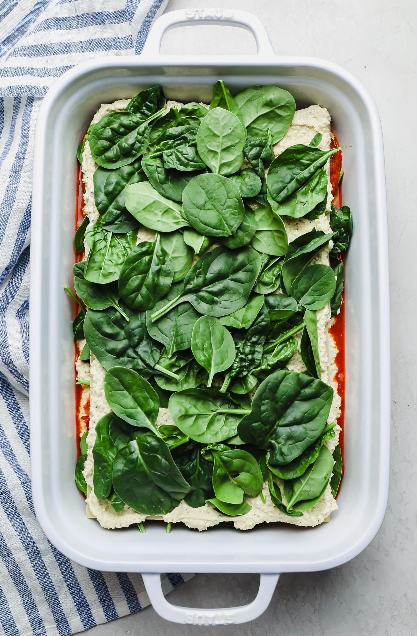 spinach on top of a layered, unbaked vegan lasagna in a white casserole dish.