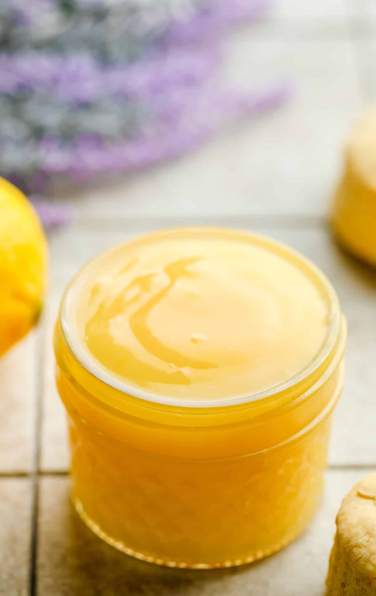small jar of vegan lemon curd with lavender in background, lemons on a grey squared board