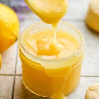 close up of lemon curd being drizzled into the jar with a white spoon on grey background