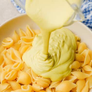 pouring yellow cashew cheese sauce over a large bowl of cooked pasta.