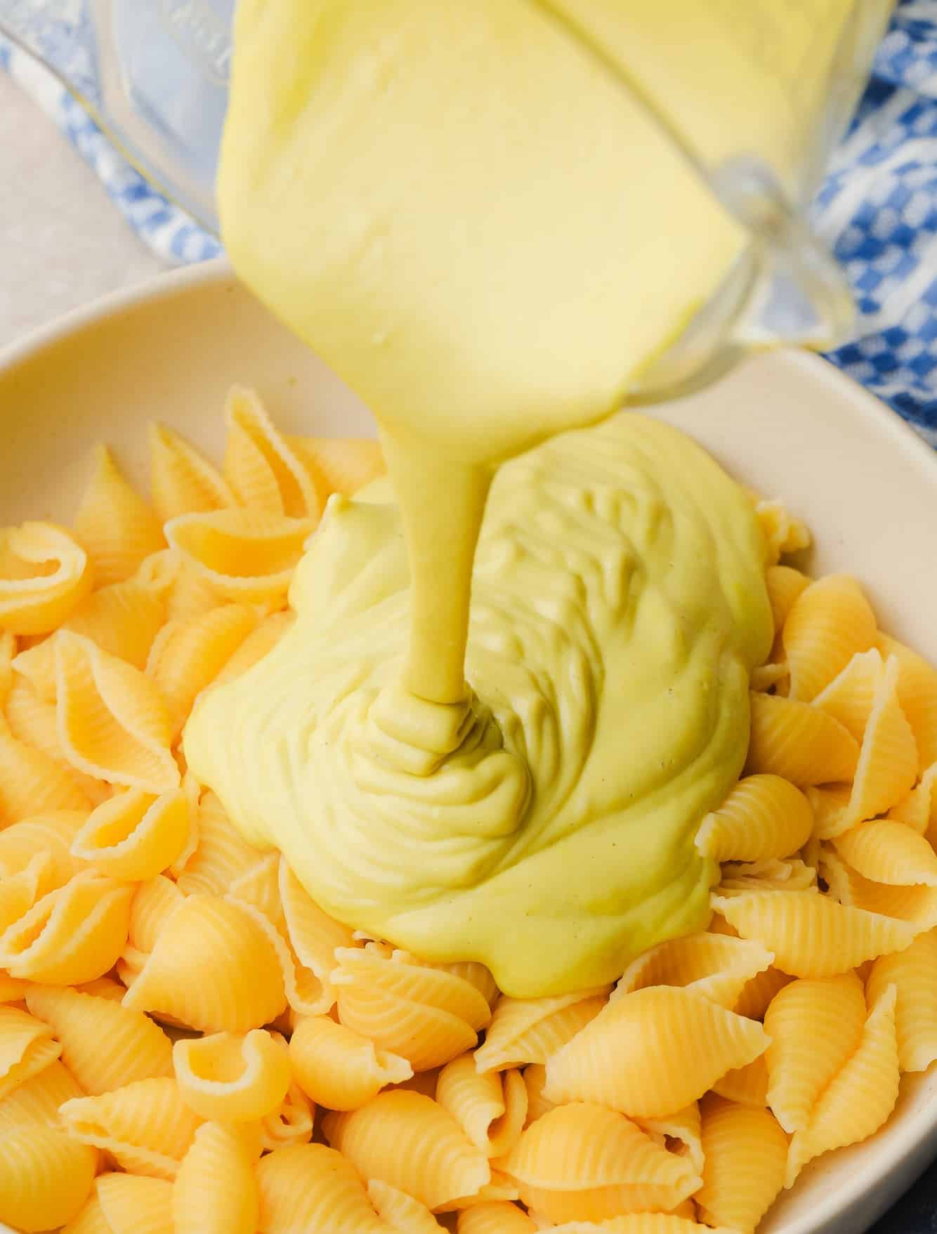 pouring yellow cashew cheese sauce over a large bowl of cooked pasta.