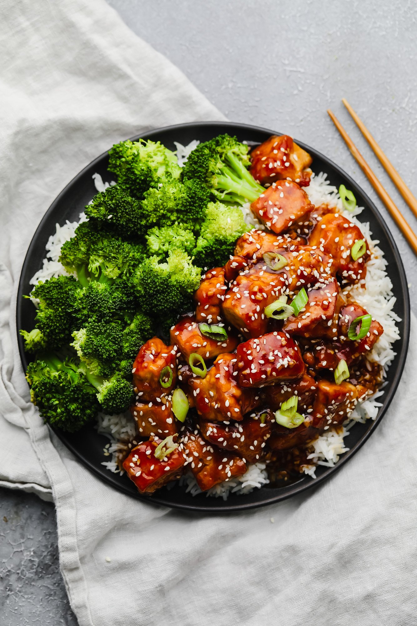 black plate with broccoli, rice and brown saucy tofu on grey background