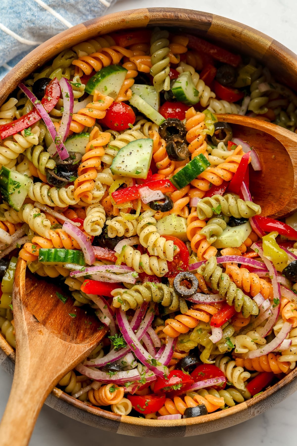 wood bowl with blue towel under it of vegan pasta salad with two wood spoons in it