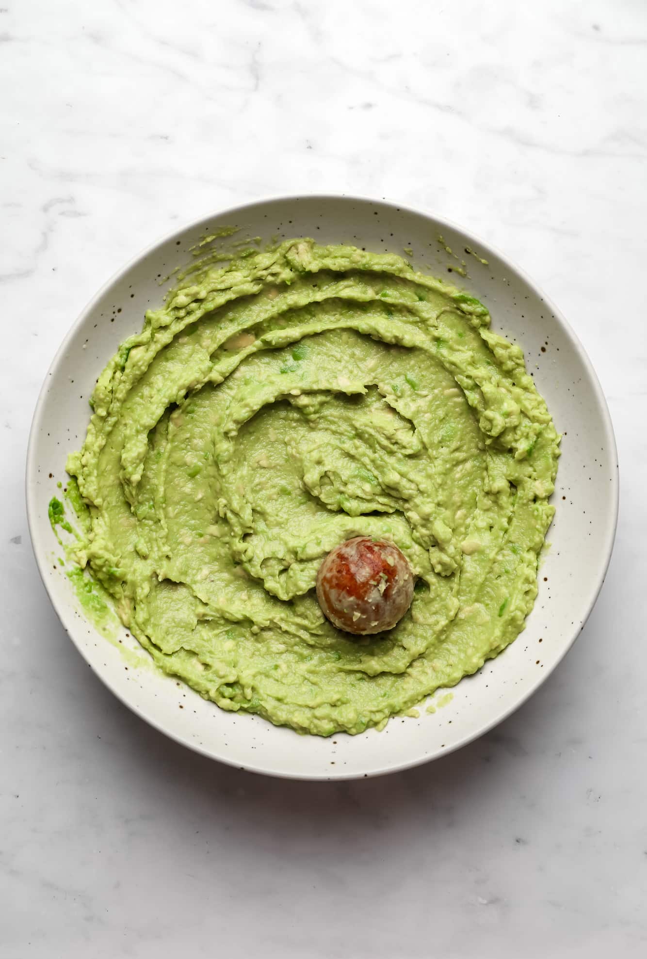smooth and green avocado spread with the avocado seed on top in a beige bowl.