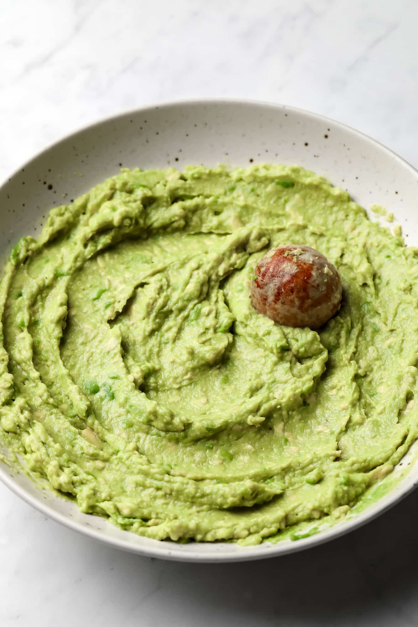 smooth and green avocado spread with the avocado seed on top in a beige bowl.