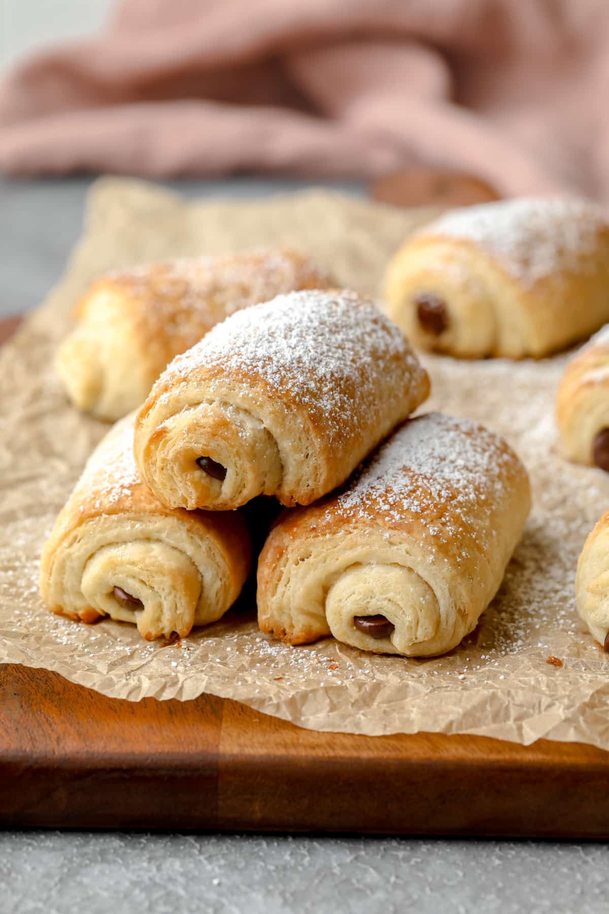 3 vegan pain au chocolat stacked on crinkly parchment paper with pink towel in background and more croissants