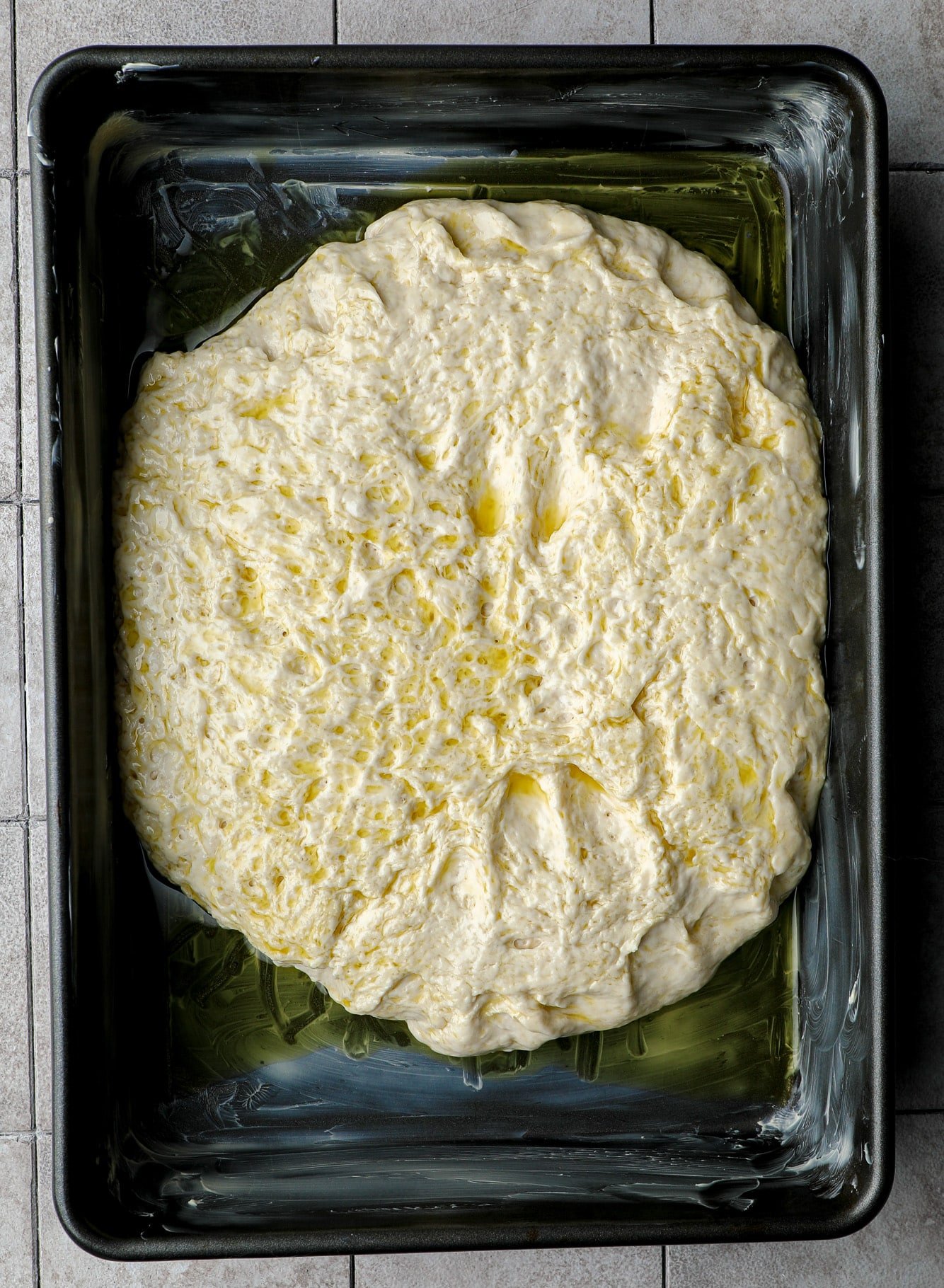 oiled focaccia dough in a large black baking pan.