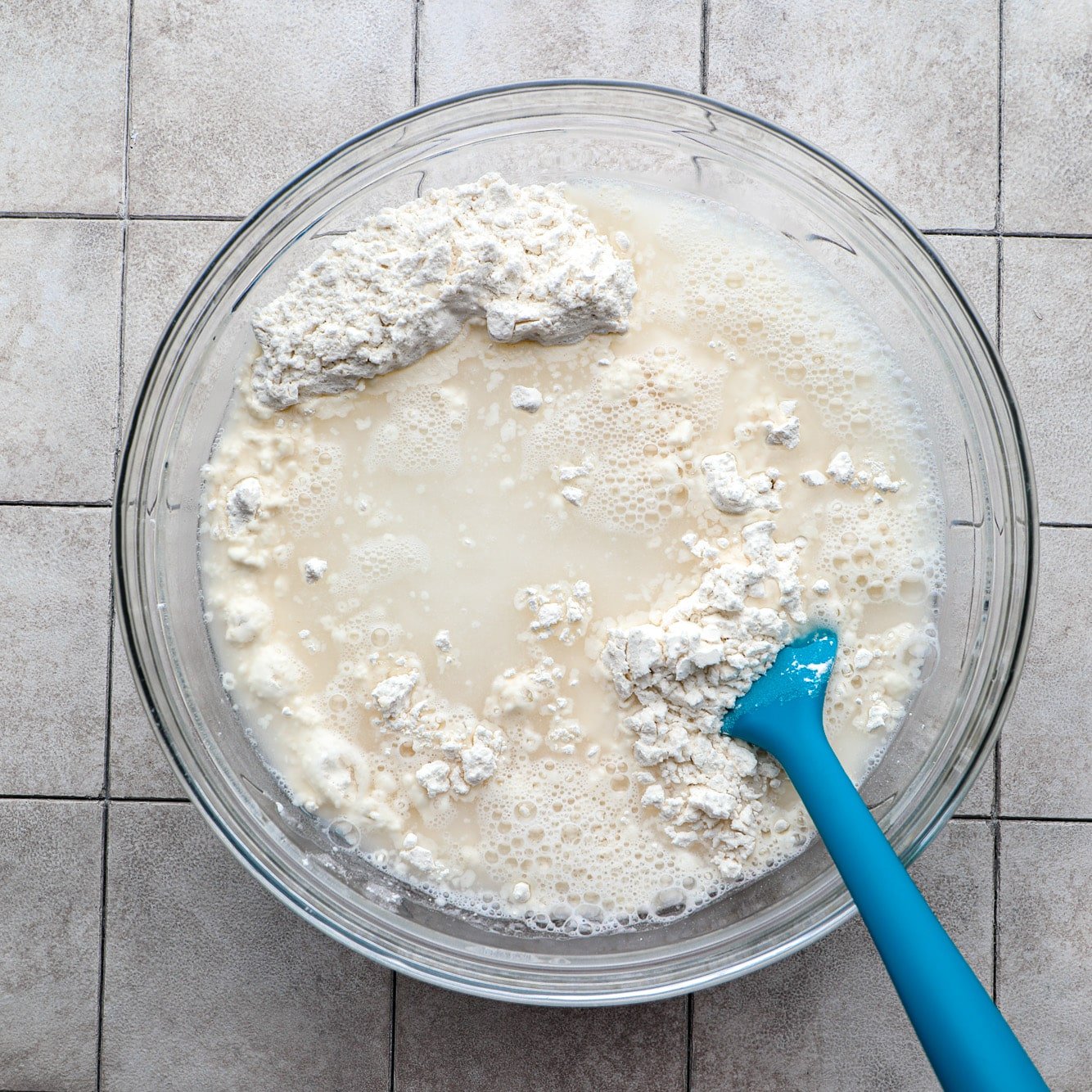 using a blue spatula to stir focaccia dough ingredients in a glass bowl.