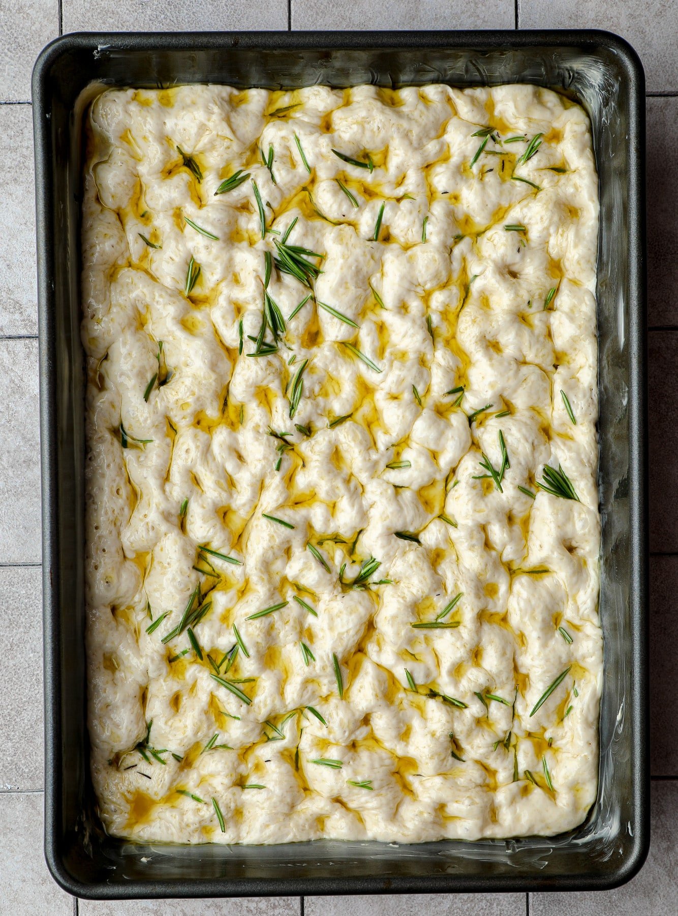 dimpled vegan focaccia dough in a black pan with oil and rosemary on top.