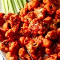 close up on buffalo cauliflower wings on a white platter with celery sticks and ranch dipping sauce on the side.