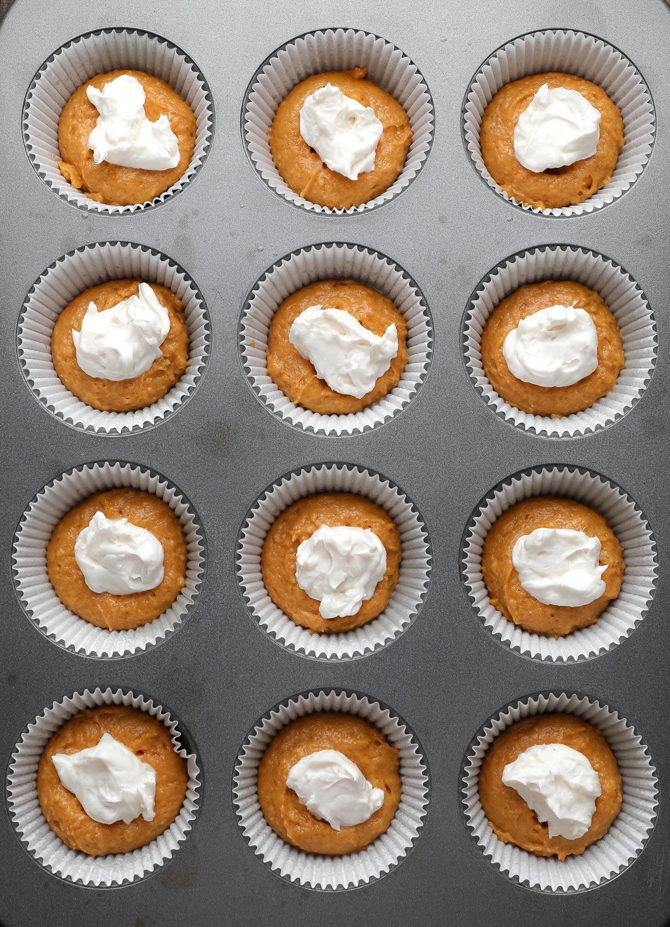 a muffin tin filled with vegan pumpkin muffin batter topped with dollops of vegan cream cheese.