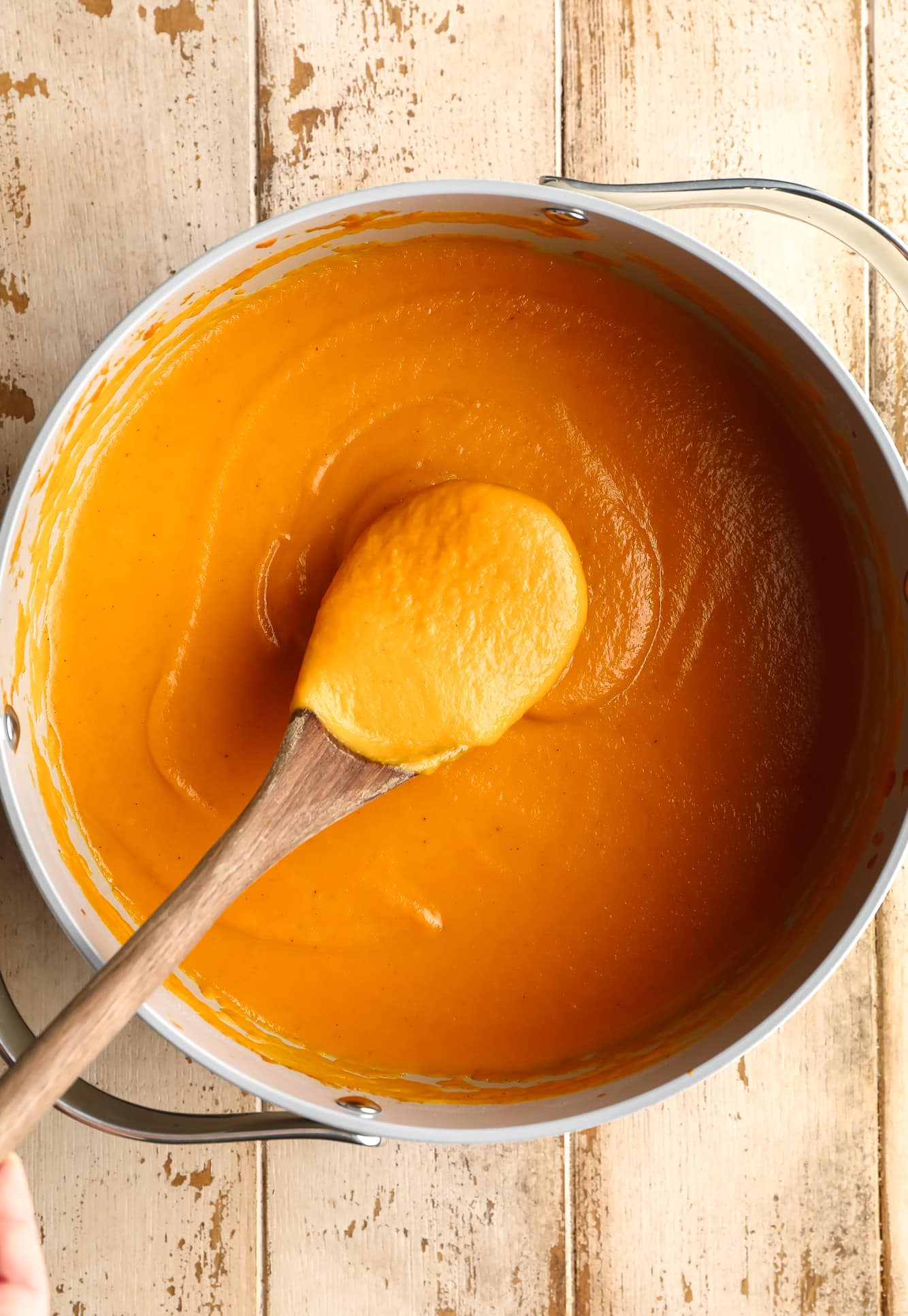 a wooden spoon lifting up a scoop of vegan pumpkin soup from a large batch in a grey pot.