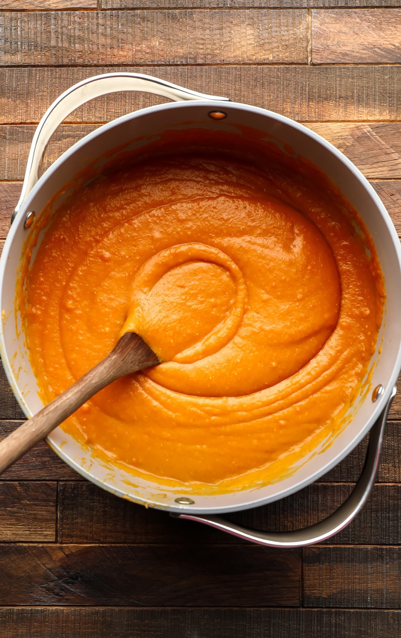 using a wooden spoon to stir creamy carrot and lentil soup in a large grey pot.