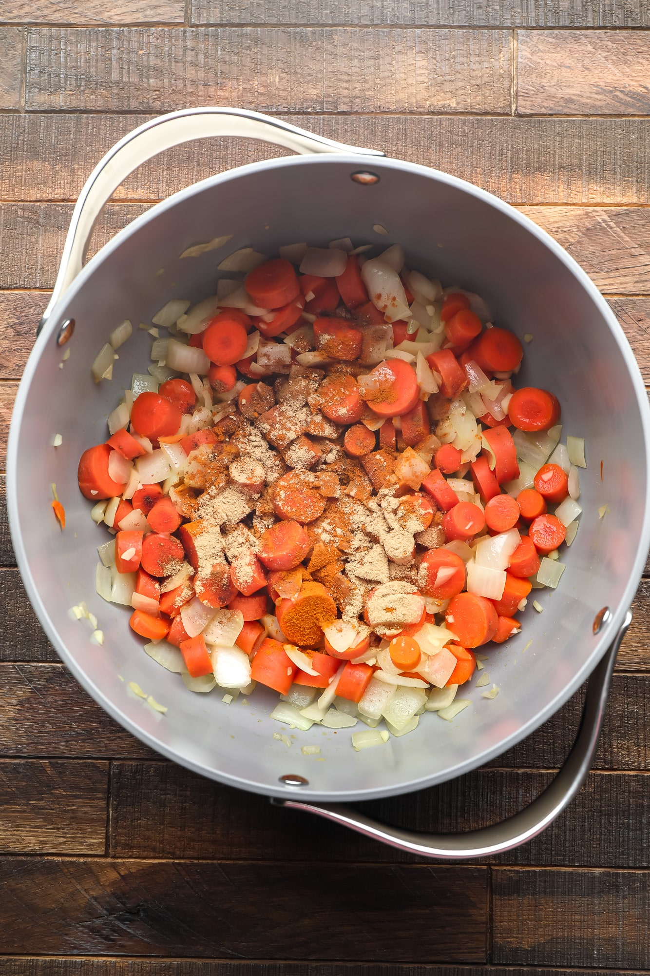 chopped carrots, onions, and garlic coated in spices in a large grey pot.